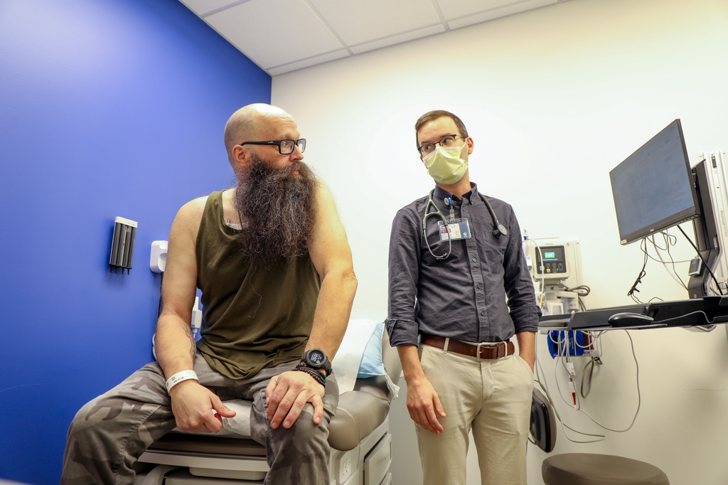 Clifford Alexander, a bearded man with a shaved head wearing camo pants and a tank top, sits on an exam table during a check-up with a physician assistant.